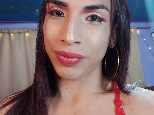 Uma selfie de brunettehot77 com batom vermelho, sombra rosa, pele brilhante, brincos de argola e um sutiã de renda vermelho, mostrando uma pequena pinta em sua bochecha..