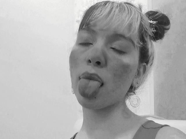 ElizabethBlizz mit zerzaustem, blondem Haar zu einem Dutt gebunden, mit geschlossenen Augen und herausgesteckter Zunge, trägt ein Top und ihr Gesicht ist mit einer dicken, weißen Maske bedeckt. Schwarz und Weiß..