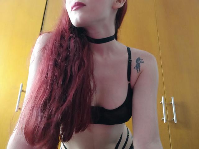 AkemiHaishi with long red hair, black choker, black bra, tattoo on right shoulder, pale skin, looking down, yellow wooden doors in background, visible straps on black underwear.