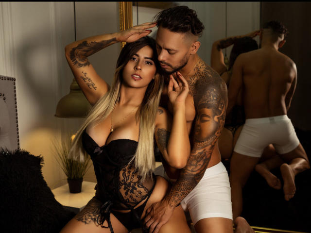 KendraWaltonJeffLuthor, met grote borsten, zwart kant lingerie en tatoeages, staat naast de getatoeëerde JeffLuthor in witte onderbroek, beiden in de reflectie van een spiegel, waarbij ze haar arm aanraakt..