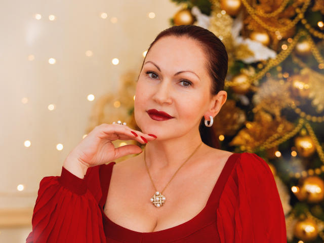 Rene69Ice with red dress, puffed sleeves, red lipstick, red nails, necklace, earrings, smiling, brown hair in bun, Christmas tree with gold ornaments in background. Boobs visible.