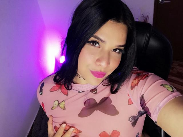 AntonelaPark with black hair, pink lipstick, and a choker, wearing a pink shirt with butterfly prints, holding her small tits, purple light in background.