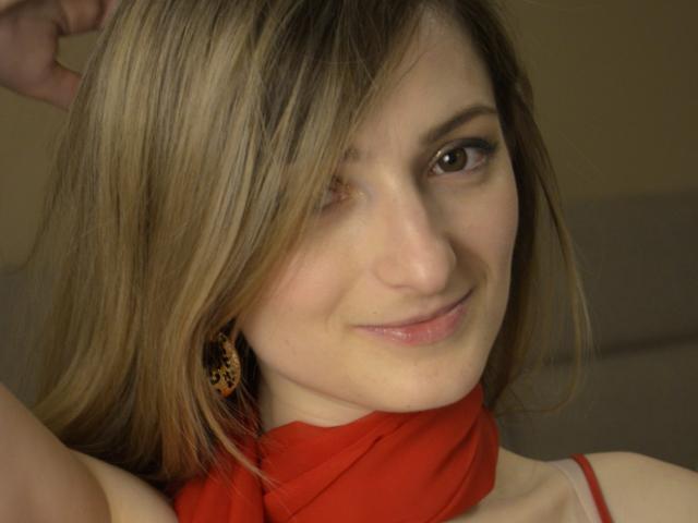AriaMermaid with blonde hair, a red scarf, and leopard earrings, smiling slightly, has one eye partially closed, and is touching her hair with her right hand.