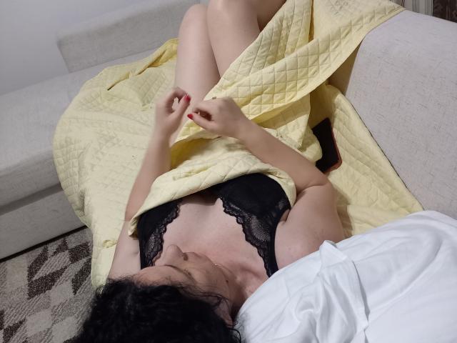 KinkyKosmina with pale skin, black lace bra, and curly dark hair, lying on a beige sofa, covering her tits with a yellow quilt, red-painted nails, white pillow.