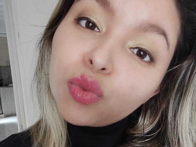 Myalewwis, con labbra rosa carnose, occhi marrone chiaro e capelli castano con riflessi biondi, indossa un maglione nero a collo alto e ha un'espressione carina, leggermente confusa..