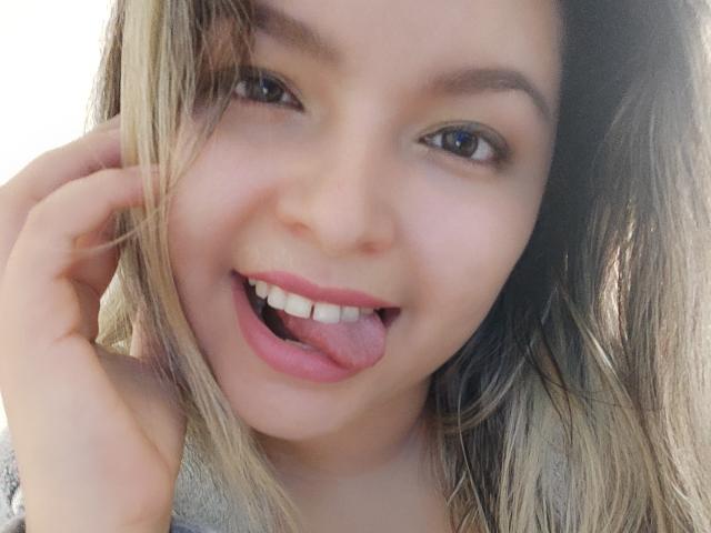 Myalewwis's face, blonde hair, blue eyes, and pink tongue out, smiling with her hand near her ear. She has fair skin, and her teeth are straight.