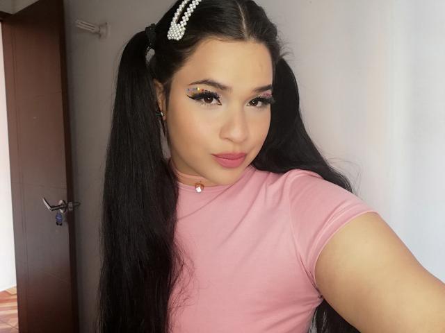 AntonelaPark with long black pigtails, wearing a pink shirt, has bold eyeliner and pink lipstick, pearl choker, and pearl hairpins, looking at the camera with a slight smile.