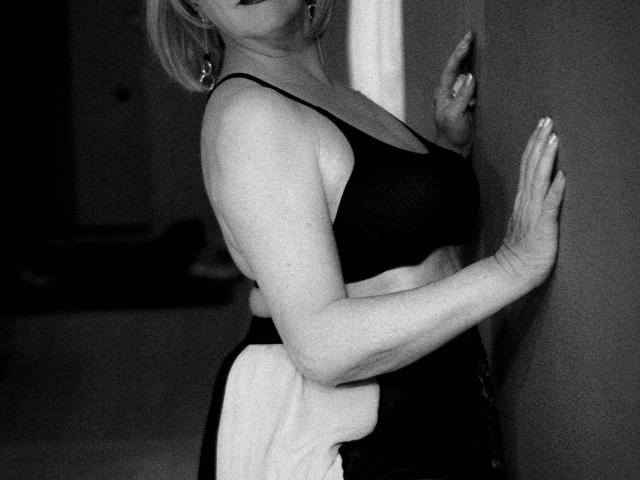 SamanthaLala, con un seno di media grandezza, indossa un reggiseno nero e una gonna bianca, è in piedi contro un muro, con la mano destra appoggiata al muro e la mano sinistra sollevata, fotografia in bianco e nero..