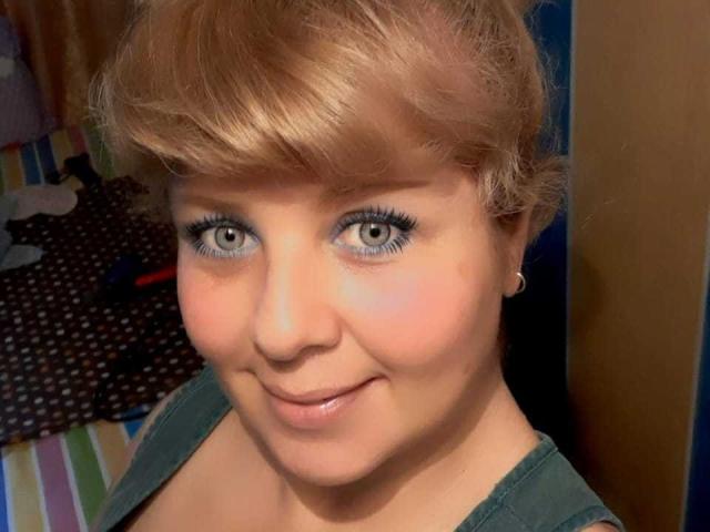 Casal latino com cabelo loiro, olhos azuis e um sorriso adorável, vestindo uma camisa verde, pequenos brincos de argola e delineador azul, olhando diretamente para a câmera..