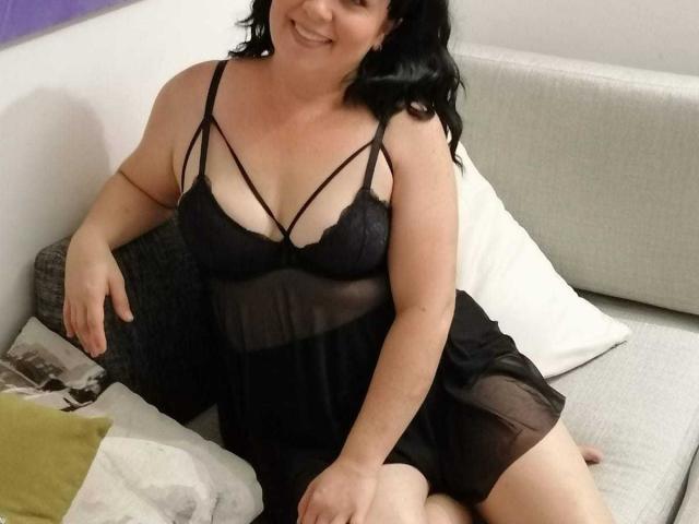 WildLatinCouple es una mujer curvilínea con lencería de encaje negro, que muestra sus senos en un vestido negro transparente, sentada en un sofá gris, sonriendo, con cabello negro y piel pálida..