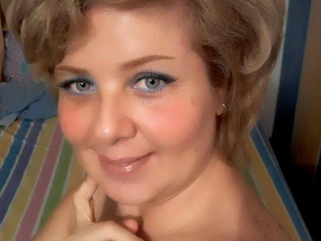 A WildLatinCouple tem cabelo loiro curto, maquiagem azul nos olhos e um leve sorriso com lábios brilhantes, tocando o queixo. Seus seios não estão visíveis, e ela está sem blusa..