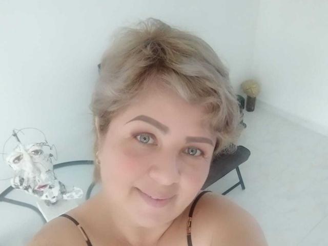 WildLatinCouple tiene el cabello rubio corto y despeinado, ojos verde claro y una ligera sonrisa, vestida con un top negro con tirantes, donde se pueden ver pequeños senos, en una habitación luminosa..