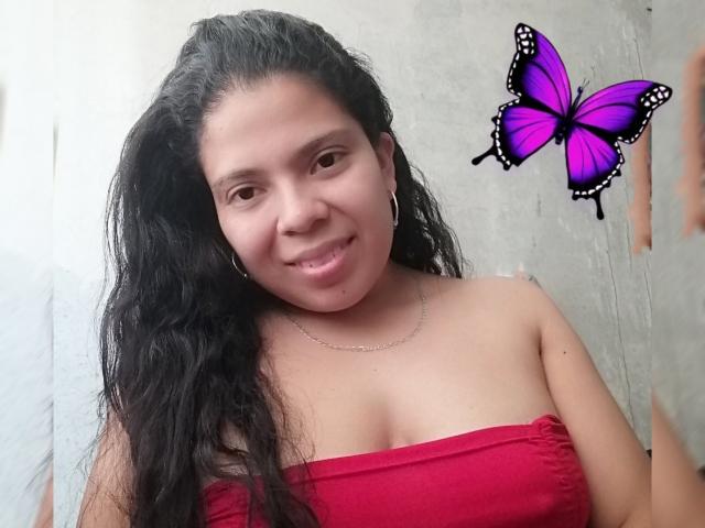 EstrellaBelle, com cabelos longos e pretos, sorrindo, vestindo um top vermelho sem alças, mostrando os seios, com um adesivo de borboleta roxa no lado direito e um colar de prata..
