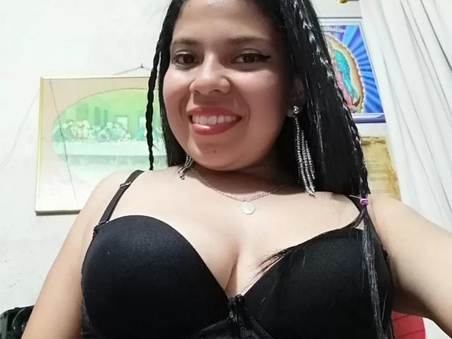 EstrellaBelle, com longas tranças pretas, sorrindo, usando um sutiã preto que mostra seus seios grandes, um colar de prata e brincos, em um selfie..