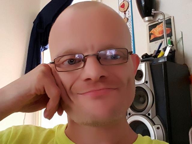 Ein kahlköpfiger CallMeHotX mit Brille, einem hellgelben Hemd und einem leichten Grinsen, der/die den Kopf in der Hand ruht. Stereos im Hintergrund..