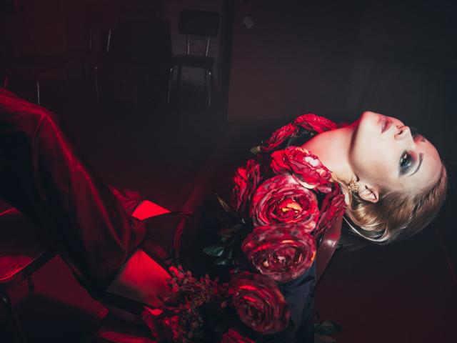 LexiexWild com rosas vermelhas cobrindo seus seios, inclinada para trás com um olhar sedutor, luz vermelha realçando sua pele clara e maquiagem escura, em um ambiente escuro e melancólico..