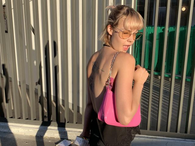 LexiexWild, con cabello rubio y gafas, lleva una blusa rosa y negra, mostrando su espalda con tatuajes, sosteniendo un bolso y de pie frente a una valla de metal iluminada por el sol..