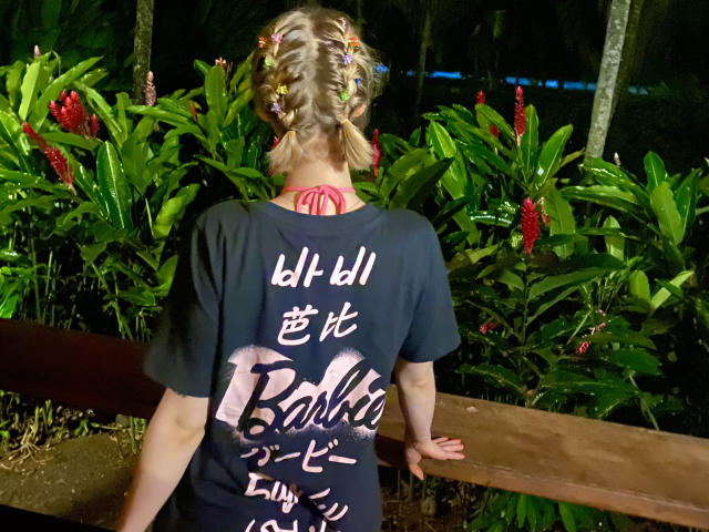LexiexWild, con el cabello trenzado, vestida con una camisa negra, está de espaldas a la cámara, de pie frente a plantas verdes con flores rojas, con las manos sobre una barandilla de madera..