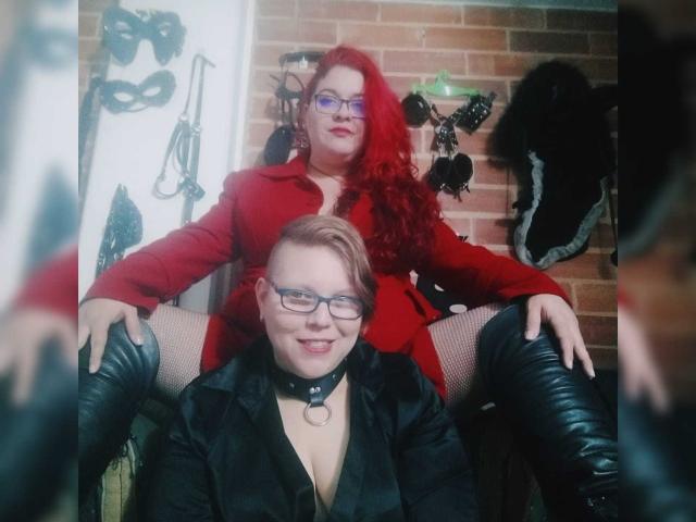 Due ragazze latine, una con i capelli rossi e gli occhiali, un vestito rosso e degli stivali, seduta in grembo all'altra, che indossa un completo di pelle nera, calze a rete e un collare..