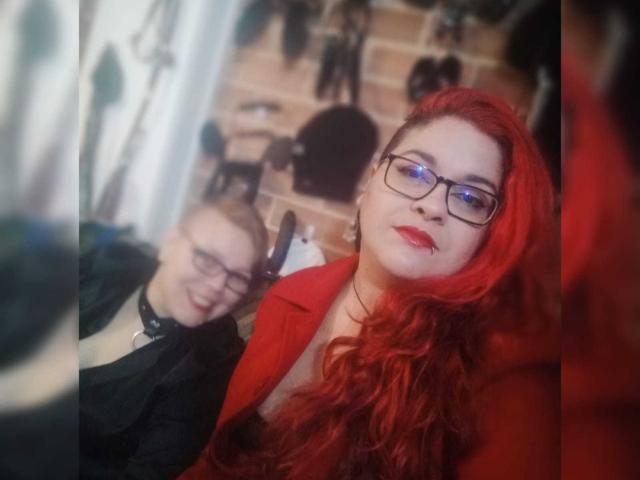 LatinoHardGirls met rood haar, een bril en rode lippenstift, die een rode jas dragen en staan naast een kale LatinoHardGuy met een bril, gekleed in het zwart. Vage achtergrond..