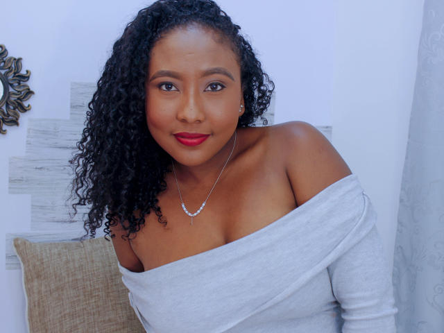 CaroldelaTorre with curly black hair, brown skin, red lipstick, off-shoulder grey top, showing tits, silver necklace, smiling at camera.