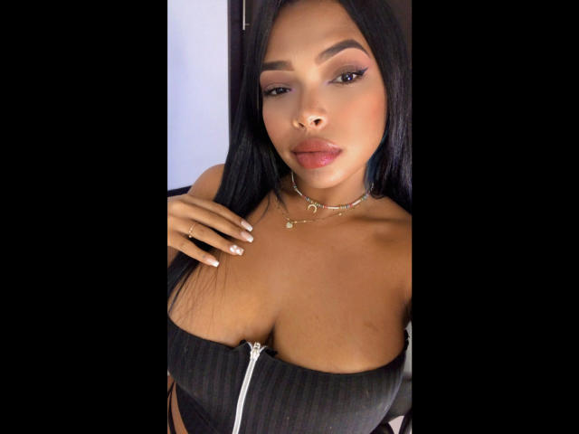 VickyGreys with tan skin, long black hair, and full lips, wearing a black ribbed top with a white zipper, and a gold necklace, showing her tits and a nose ring.