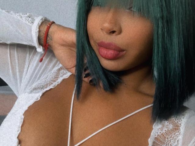 VickyGreys with teal hair, full lips, and white lace top, showing her tits, arm raised, red bracelet, and white necklace, looking down.