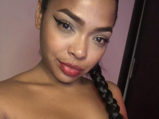 VickyGreys com pele escura, batom vermelho, cabelo trançado preto, sobrancelhas grossas e delineador, sorrindo levemente, mostrando um piercing no nariz, sem blusa, em um quarto com pouca luz..