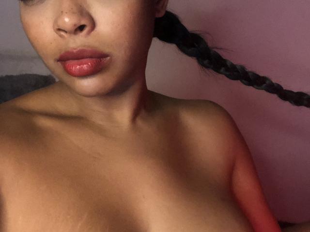 VickyGreys has brown skin, glossy red lips, and a braided ponytail, showing her bare shoulders and small tits against a pinkish background. Her nose has a small freckle.