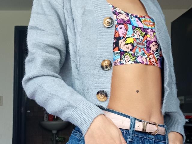 El abdomen de Yukio, con un busto pequeño, lleva una camiseta corta de colores con un estampado de cómic, un cárdigan azul claro con botones y jeans azules con un cinturón rosa. Se puede ver el ombligo..