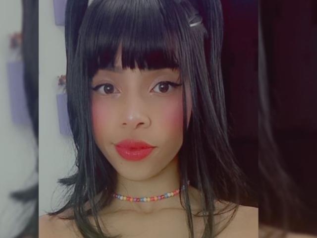 Yukio tem rabos de cavalo pretos, franja e um colar colorido de contas. Ela está usando batom vermelho e tem blush rosa nas bochechas. Seus olhos são escuros e ela está sorrindo..