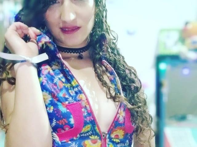 KingAndQueeSweet, com cabelos castanhos cacheados, vestindo um vestido colorido com estampa floral e zíper, zíper rosa, colar preto e fita branca no pulso, exibindo um sorriso adorável com batom rosa..