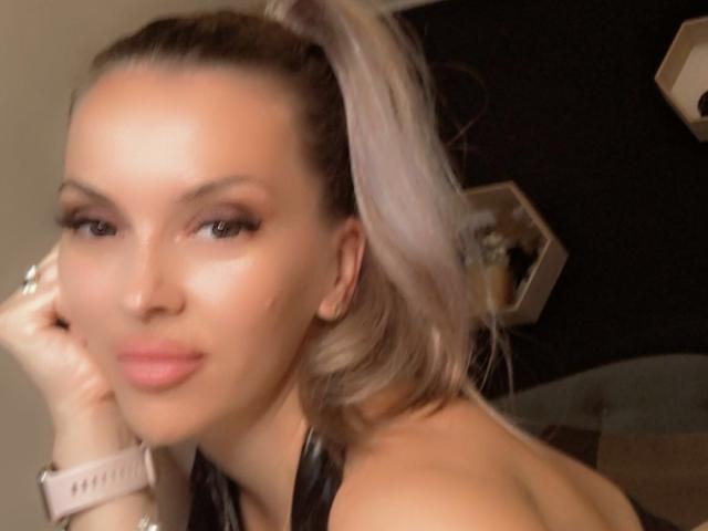 SophieDeee with blonde ponytail, pink lips, and shiny skin, wearing a pink watch, resting her chin on her hand, showing her tits and a slight smile.