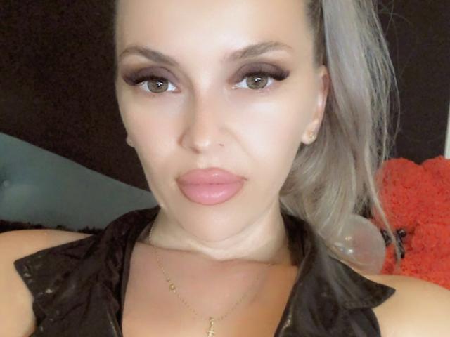 SophieDeee has puffy pink lips, thick black eyeliner, and light green eyes. She's wearing a black sleeveless top, and has a gold necklace. Her gray hair is in a ponytail.