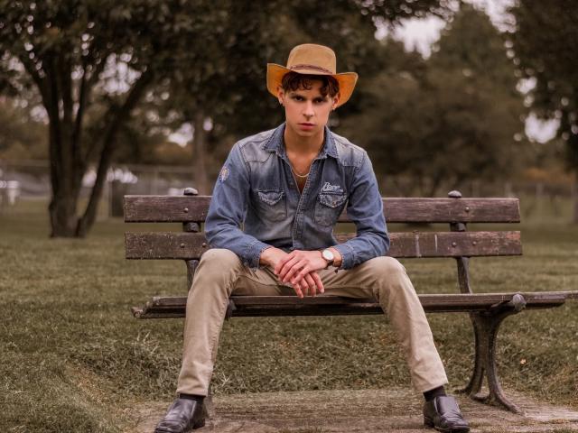 Logan Evans sitzt auf einer hölzernen Bank, trägt einen beigen Hut, ein blaues Jeanshemd, beigefarbene Hosen und schwarze Schuhe, hat die Hände auf den Knien und wirkt ernst..