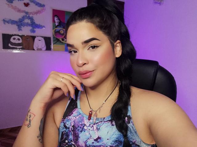 AntonelaPark with dark hair in a braid, wearing a blue galaxy tank top, pink lipstick, and a red stone necklace, resting her chin on her hand, showing a tattoo.