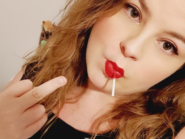 MaitresseAnna, com cabelos castanhos cacheados, batom vermelho e um pirulito na boca, apontando para os lábios com o dedo indicador. Ela está vestindo uma blusa preta..