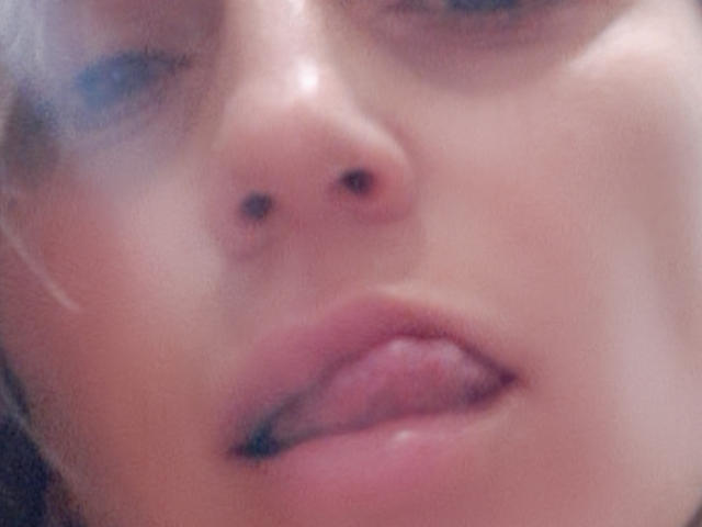 AliaAnita's face, with blurred pinkish skin, shows her nose, slightly parted pink lips with tongue touching upper lip, and blue eyes, creating a soft, sensual, close-up shot.