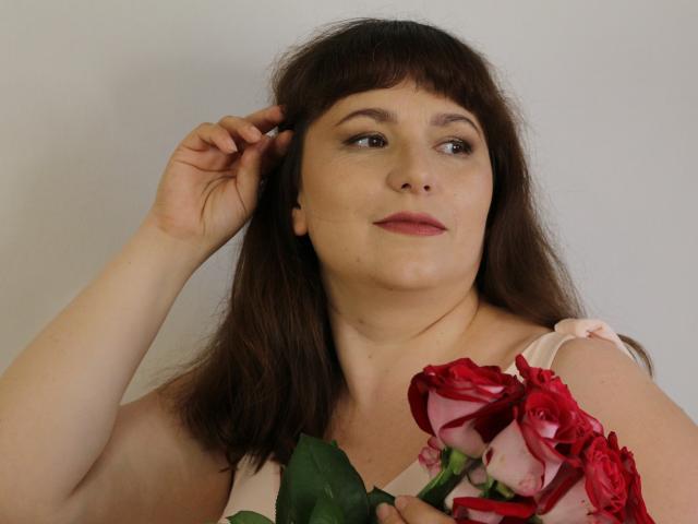 AliceInks with brown hair, holding red and pink roses, has a hand in her hair, looking slightly to the side with a calm expression, wearing a pink sleeveless top.