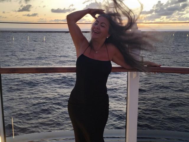 EvaRodinson, avec ses longs cheveux bruns, vêtue d'une robe noire à encolure dégagée, les bras levés, souriante, debout sur un bateau, l'océan en arrière-plan, coucher de soleil, nuages, rambarde en bois, eau, détendue, heureuse..