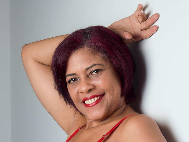ErikaSpmt with red hair, smiling, wearing red lingerie, arm raised, showing off her tits, has red lipstick, and is looking at the camera.