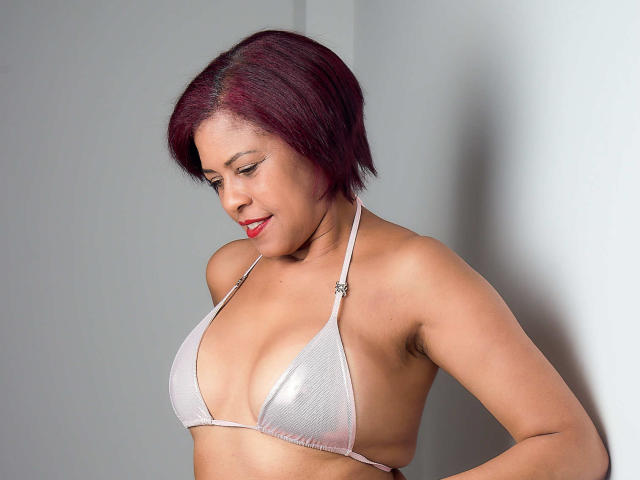 ErikaSpmt with short red-brown hair, wearing a shiny silver bikini top, showing her tits, armpit visible, looking down with a slight smile.