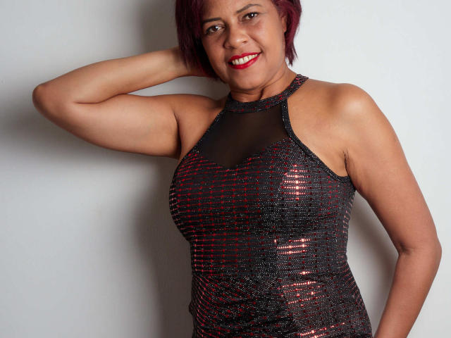 ErikaSpmt has red hair, red lipstick, and a shiny black and red sequined dress, showing her tits and smiling with her arm raised. She's standing against a plain wall.