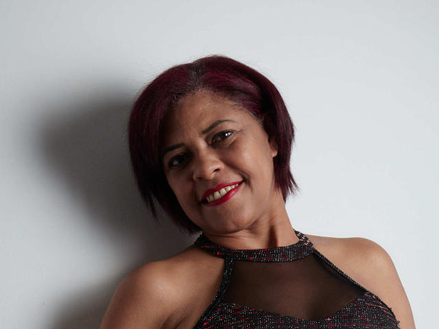 ErikaSpmt, com cabelo roxo curto, sorrindo, vestindo uma blusa preta com alças finas e brilhantes, mostrando seus ombros nus e usando batom vermelho. Ela tem a pele marrom média e está olhando para a câmera..