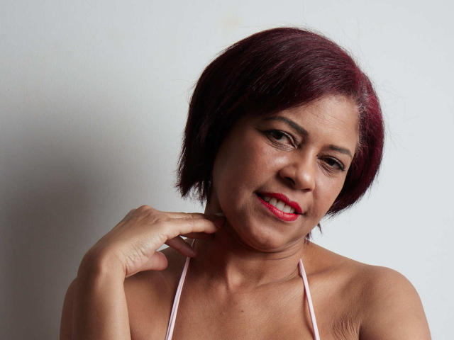 ErikaSpmt hat kurze, rote Haare, hellbraune Haut und roten Lippenstift. Sie lächelt und hält ihre Hand in der Nähe ihres Halses, sie trägt ein pinkes Top mit Halter. Ihre Brüste sind klein..