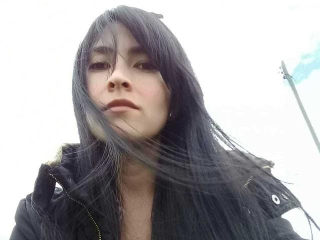 JainaRogers a de longs cheveux noirs, elle porte une veste noire, et une légère brise souffle dans ses cheveux, avec une expression neutre sur son visage..