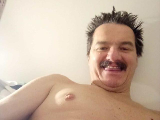AngeloBestia est un homme torse nu, avec une moustache hérissée et un sourire heureux, montrant un petit mamelon sur sa poitrine pâle et poilue. Il a des cheveux noirs et hérissés..