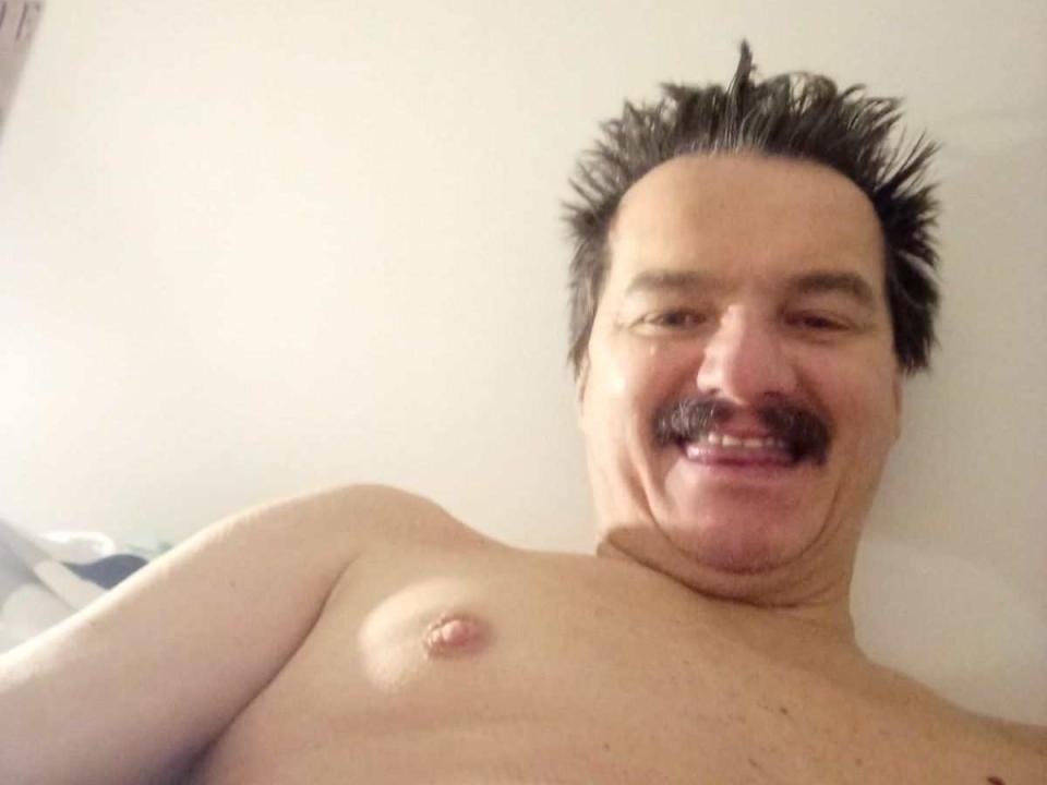 AngeloBestia ist ein Mann ohne Hemd, mit einem stacheligen Schnurrbart und einem fröhlichen Lächeln, der eine kleine Brustwarze auf seiner blassen, behaarten Brust zeigt. Er hat dunkles, stacheliges Haar..