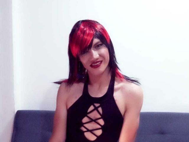 LunaGransTs with red and black hair, smiling, wearing a black halter dress with a criss-cross front, sitting on a gray couch. Boobs visible through dress.