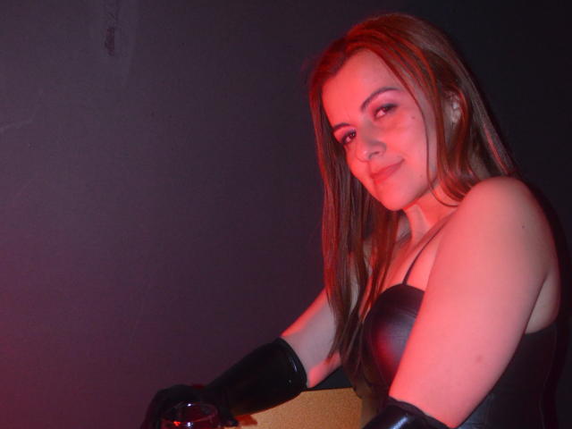 PerlaDivaio with long brown hair, smiling slightly, wearing a black leather strap top and black gloves, red lighting on her face, holding a yellow object.